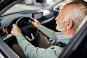 Rear view of old driver with hands on steering wheel driving a car in traffic on city street.