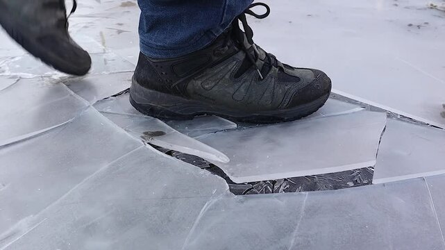Close-up of a man wearing winter and hiking boots stepping on fragile ice, shattering it like glass. The crisp sound of breaking ice enhances the immersive visual and auditory experience.