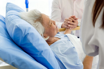 An elderly woman lies in a hospital bed, receiving comfort and support from a doctor and a family...