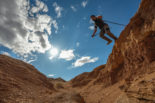 A pioneer traveler with trekking poles in his hands makes a jump in a canyon located in the Gobbi Desert in Mongolia.