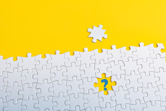 A white jigsaw puzzle on yellow background. A piece is missing in the center, with a blue question mark, a puzzle piece placed above, symbolizing problem-solving or searching for the right fit.