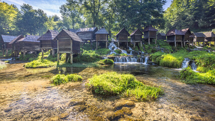 The Mlincici wooden watermills at Pliva river near Jajce town in Bosnia and Herzegovina, Europe. Scenic landscape of wooden huts elevated above a stream, surrounded by lush green trees.