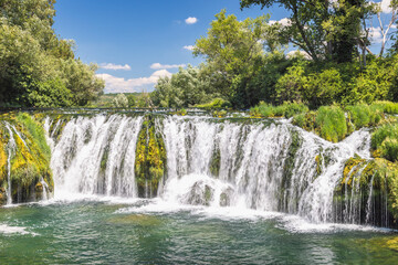Fototapeta premium Kocusa waterfall in village Veljaci, Bosnia and Herzegovina, Europe. Picturesque waterfall cascading over mossy rocks under a bright blue sky surrounded by lush green trees on a sunny day.