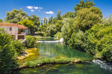 Kocusa waterfall in village Veljaci, Bosnia and Herzegovina, Europe. Riverside house nestled among lush greenery, with clear turquoise water and a small waterfall under blue sky with scattered clouds.