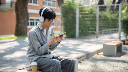 Obraz premium Smiling asian man with glasses is watching video or playing game on his phone, sitting on the bench.