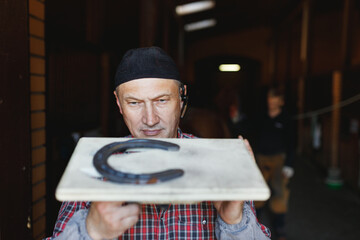 European busy blacksmith inspects horseshoe made for horse in stable