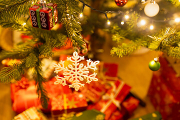 A warm, glowing scene featuring a decorated Christmas tree with a white snowflake ornament and twinkling string lights above a pile of red and green wrapped gifts. 