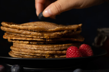 Female hand placing blueberries on stacks of pancakes made with whole wheat flour, along with raspberries on a black plate against a dark background. Homemade food, Dark food