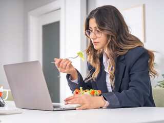 Dieting business woman eating bowl of salad in lunch break at office using laptop working while eating bowl of salad, fresh vegetarian snack. Side portrait view focused office worker employee.