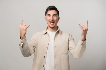 Let's rock it. Handsome brunette man in his 30s makes a cool rock sign with both hands, flashing a wide smile while looking into the camera. Trendy beige overshirt adds a modern touch