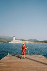 Fototapeta premium Little girl with a pink soft toy stands on the pier looking at the lighthouse. Back view