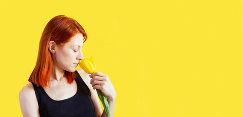 girl with red hair on a yellow background with a tulip in her hands, portrait of a young woman inhaling the fragrance of a yellow flower, place for text