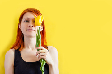 girl with red hair on a yellow background with a tulip in her hands, portrait of young woman smiling and looking at camera, space for text