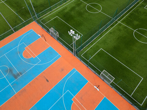 Aerial view of outdoor basketball and soccer courts with vibrant blue, orange, and green colors. The sports facility is empty, with clear markings and fences, creating a dynamic geometric composition