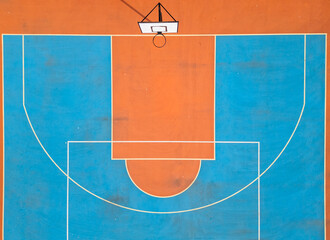 Aerial view of a basketball hoop and colorful court with vibrant orange and blue sections. The symmetrical layout and bold colors create a striking sports-themed composition