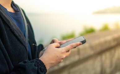 person holding smartphone outdoors near waterfront, wearing dark jacket, with blurred background of water and greenery, evoking calm and focused mood