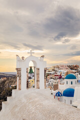 Scenic view of Oia, Santorini, Greece, featuring the iconic three domes viewpoint, colorful houses, and the Aegean Sea. Vertical shot with a cloudy sky, perfect for honeymoon or wedding destination