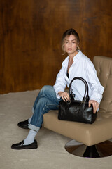 A girl in a white shirt blue jeans and loafers is posing with a black bag.