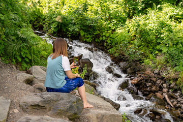Woman sitting barefoot on rocks taking photo of flowing stream with her smartphone. Surrounded by lush green foliage, she is immersed in the natural setting, walking and hiking