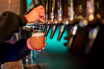 A bartender pours fresh draft beer into a glass at a bar. Focus on the hand, beer tap, and foam, with a blurred background for ambiance.