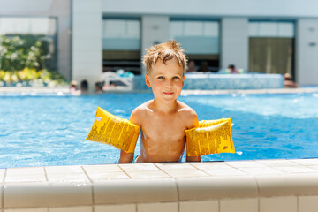 Portrait cute adorable little caucasian blond boy kid wear inflatable armband sit poolside indoor...