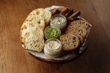 Assorted artisan bread slices with three types of butter in glass jars on a rustic wooden table. A delicious selection of fresh bakery and butter for breakfast or snack