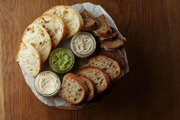 Assorted artisan bread slices with three types of butter in glass jars on a rustic wooden table. A delicious selection of fresh bakery and butter for breakfast or snack. top view