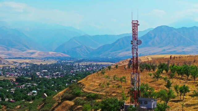 Drone footage showing a mountain village near with a telecom tower on a dry hill. The drone flying slowly in an arc around the tower, creating a parallax effect with distant mountains in the