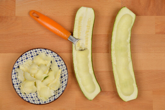 A top-down shot shows two halves of a zucchini being scooped with a melon baller, with the scooped flesh in a small bowl, on a wooden cutting board