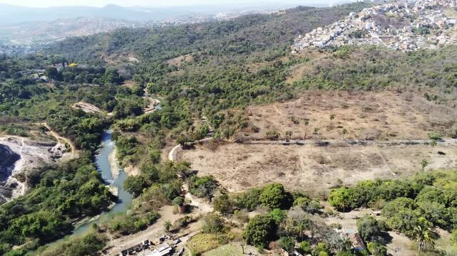 Aerial view of Izidora region in Belo Horizonte, Brazil, showcasing lush natural vegetation, farmland, winding roads, and a flowing creek amidst hills under clear skies. Nature meets urban fringe.