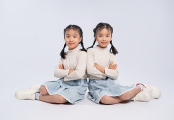 Portrait of two young identical twin sisters sitting back-to-back, with arms crossed, looking at...
