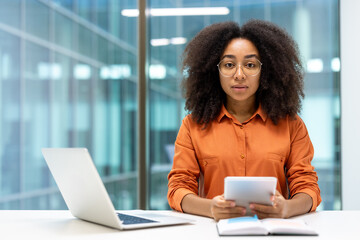 Portrait of a serious confident woman with a tablet computer in her hands. Businesswoman looks thoughtfully at the camera, works in the middle of the office, sits at a desk with a laptop