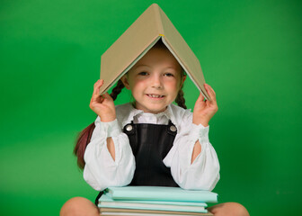 A cheerful girl with two braids holds a book over her head like a roof and smiles. She is wearing a...