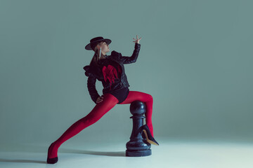 Elongated stance of woman in avant-garde outfit and red tights interacts with giant chess piece....