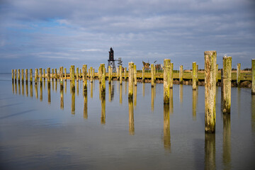 Holzpfähle in ruhigem Wasser mit bewölktem Himmel und entfernter Industriestruktur.