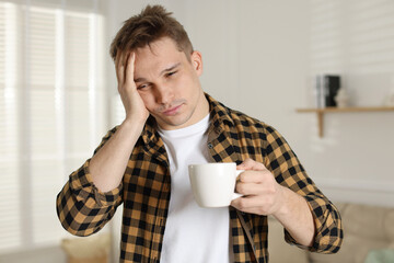 Unhappy young man with cup of coffee suffering from hangover at home