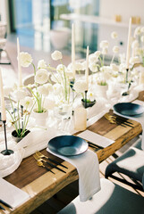 Black plates stand on a wooden table near white bouquets of flowers and candles on a narrow tablecloth