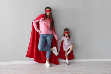 Fototapeta premium Mother and her cute little daughter wearing superhero costumes near gray wall indoors