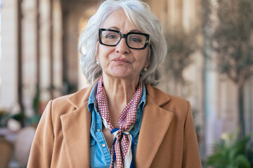 Mature woman with white hair serious at the camera. Urban setting. Elegant brown coat. Street...