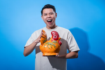 Excited young Asian man in a cream t-shirt holding a colorful ceramic rooster statue and smiling with wide expression, standing against a solid blue background, showcasing culture and creativity