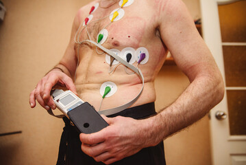 Man undergoing ECG/EKG cardiac monitoring at home with electrode patches on chest. Medical heart rhythm monitoring with electrocardiogram sensors. Holter monitor testing for heart health assessment.