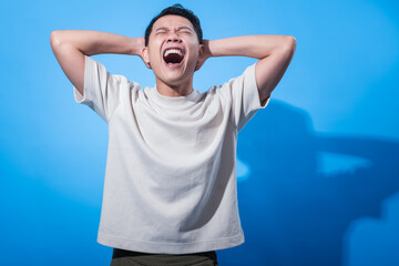 Asian man in a plain white t-shirt pulls his hair in frustration with both hands while screaming,...