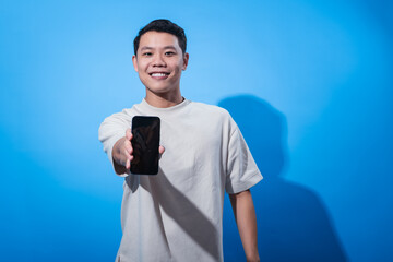 Asian man smiling confidently while holding a smartphone forward toward the camera, as if offering or presenting it. He stands against a solid blue background in a bright studio setting