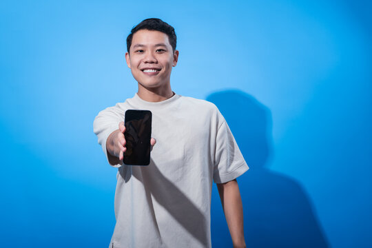 Asian man smiling confidently while holding a smartphone forward toward the camera, as if offering or presenting it. He stands against a solid blue background in a bright studio setting
