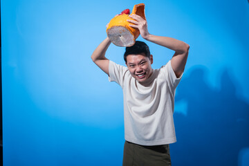 Angry young Asian man in a cream t-shirt lifting a large colorful ceramic rooster above his head with a furious expression, posing in front of a plain blue background with dramatic shadows
