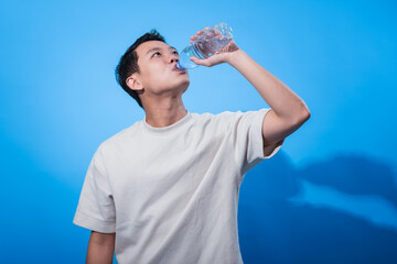 Young Asian man in a plain cream t-shirt drinks from a clear plastic water bottle while looking up, standing against a solid blue background, promoting hydration and a healthy lifestyle