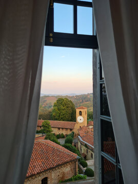 The charming countryside village of Bardassano emerges at sunset, framed by a window's soft curtains. The clock tower stands tall amidst the rustic buildings.