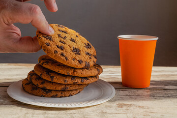 A hand carefully takes a large chocolate cookie from a stack on a white plate, symbolizing a quick breakfast, calorie-rich food, and simple pleasures. 