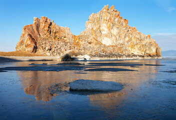 Obraz premium Spring morning on Baikal Lake. View of the famous Shamanka rock - a natural landmark of Olkhon Island in the rays of the rising sun. The bay is covered with a thin crust of blue ice. Spring landscape