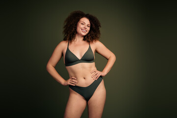 Obraz premium Confident young woman in green sportswear showcasing body positivity and natural beauty against a minimalist khaki background.